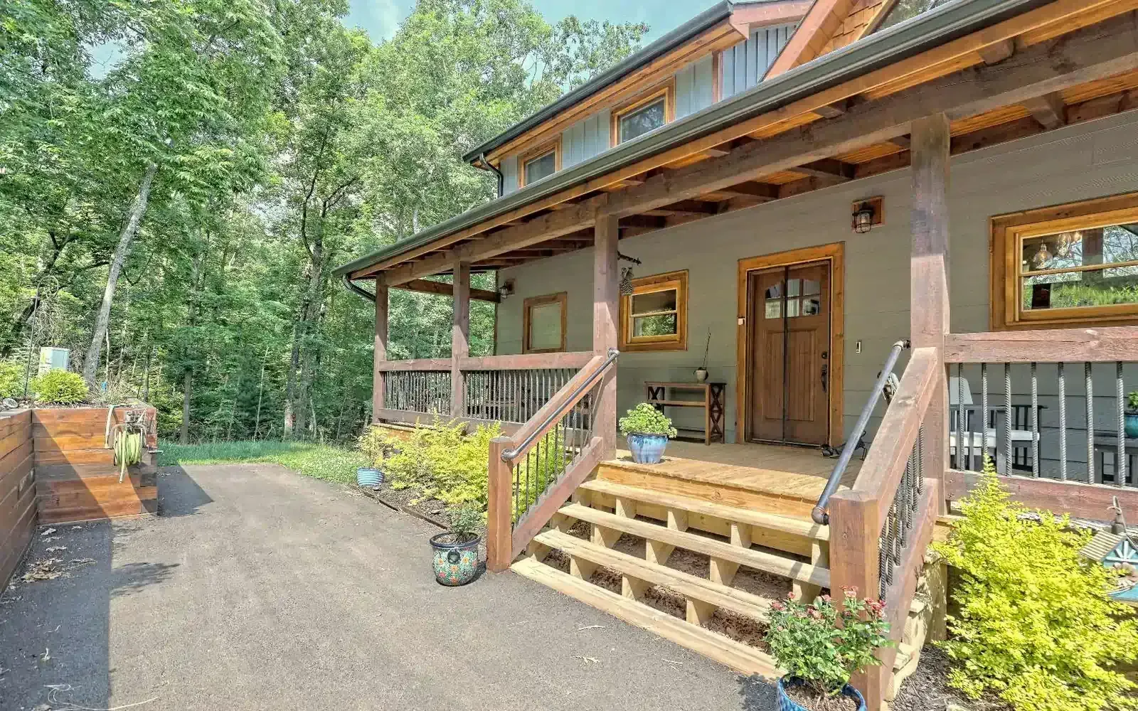 Rustic log cabin porch with wooden steps, railing, and potted plants beside a gravel driveway in a wooded setting