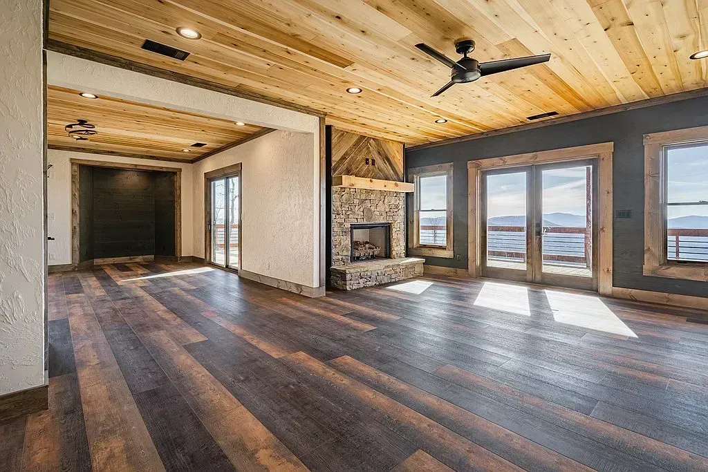 Spacious living room with wood ceiling, dark floors, stone fireplace, and large windows overlooking mountains