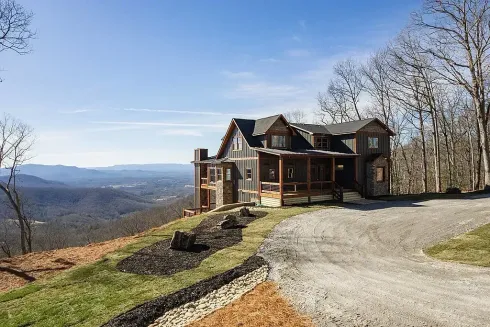 Large rustic house on a hilltop with a gravel driveway, bare trees, and a distant mountain view under a blue sky