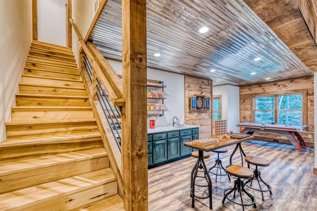 Rustic loft interior with wooden staircase, exposed beams, and a small table beside large windows.