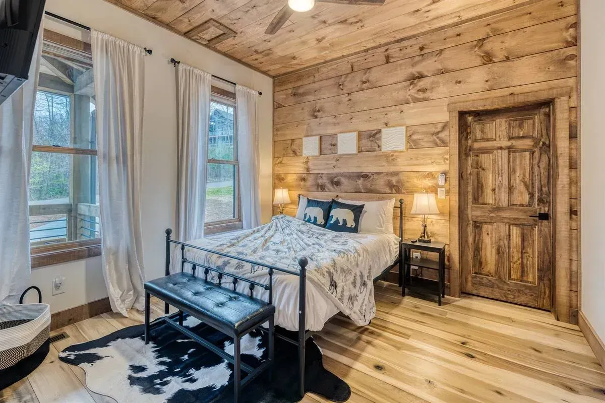 Rustic bedroom with wood-paneled walls, bed, bench, windows, and a wooden door
