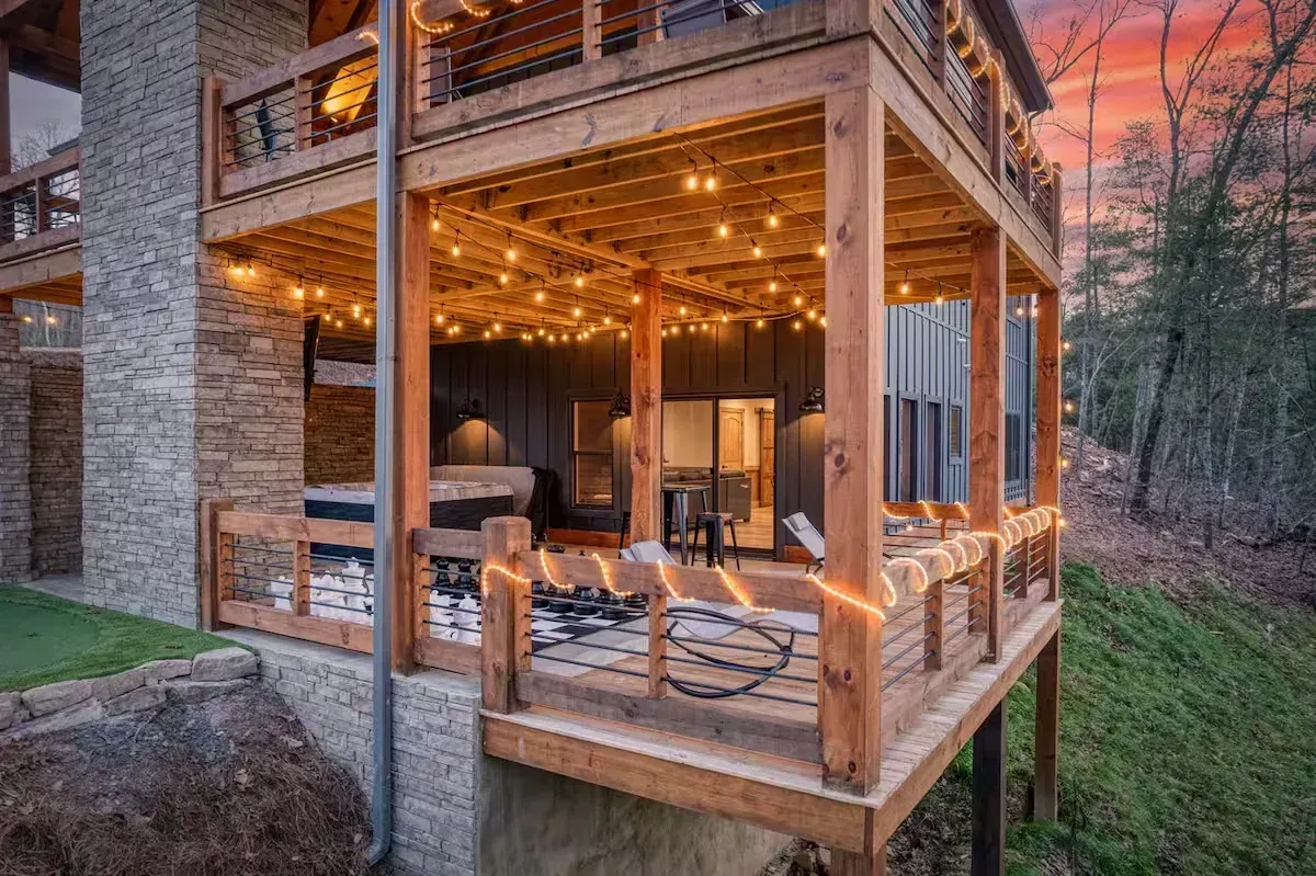 Warm, lit wooden porch beside a stone house at dusk, with seating and a forest view.