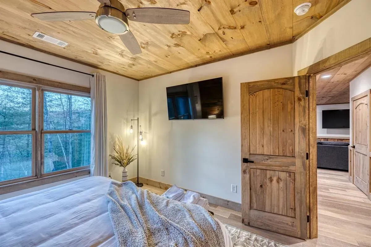 Cozy bedroom with wood ceiling, large windows, wall TV, bed, and open door to kitchen.