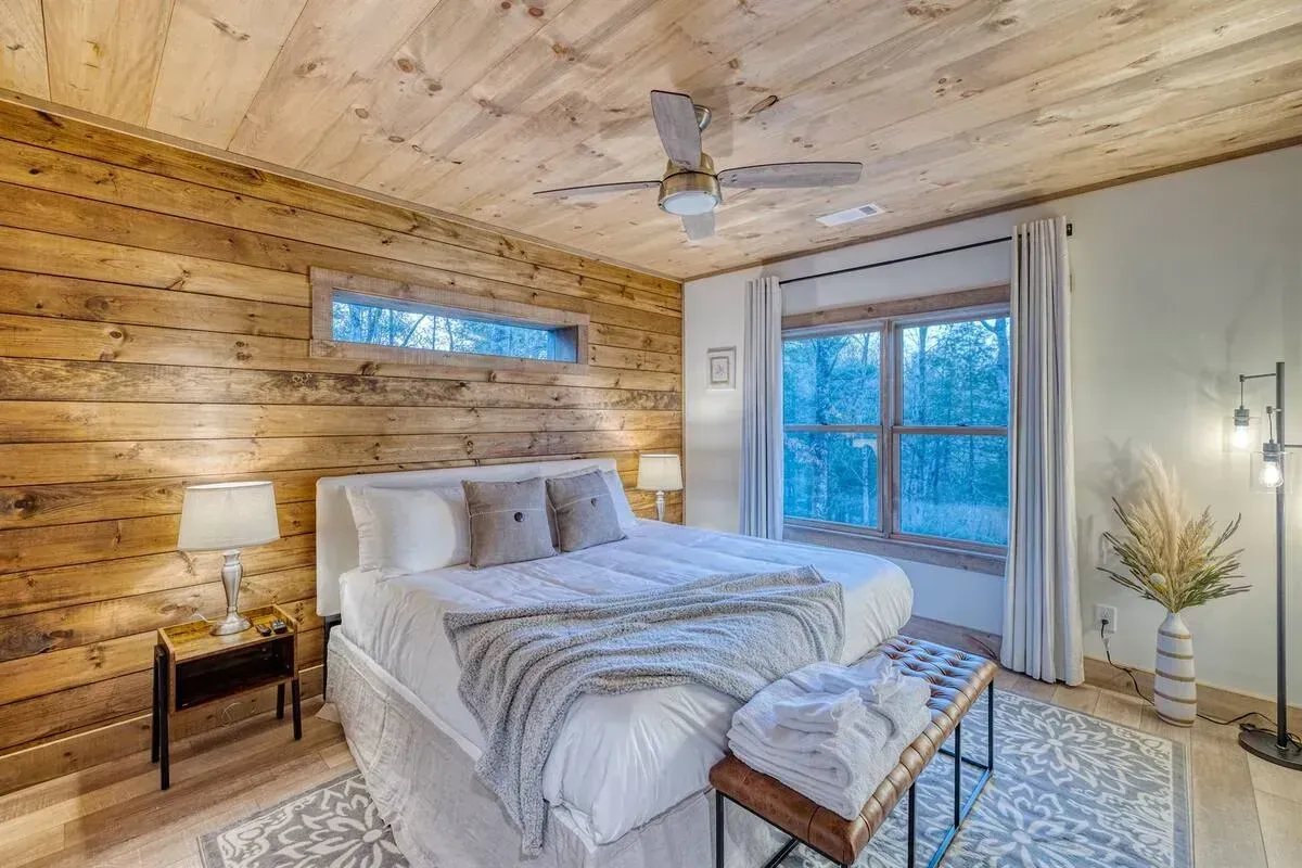 Cozy wood-paneled bedroom with a bed, bench, lamp, and large window overlooking snowy trees