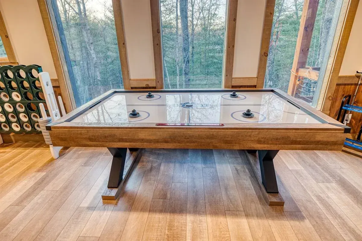 Wooden shuffleboard table centered in a bright room with large windows and trees outside.