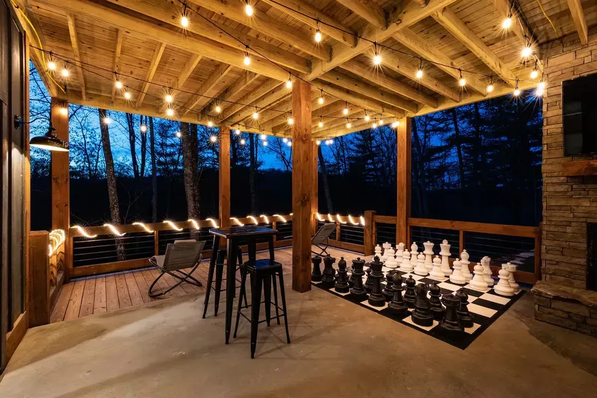 Covered outdoor chess patio with string lights, stools, and a large chessboard at dusk