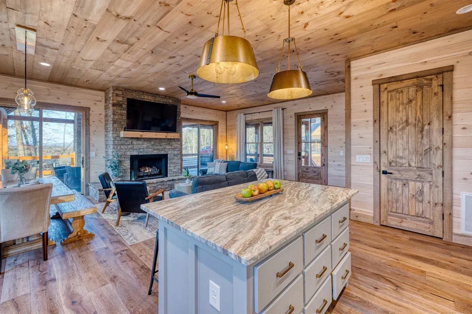 Rustic kitchen and living area with wood walls, stone fireplace, island, and large windows