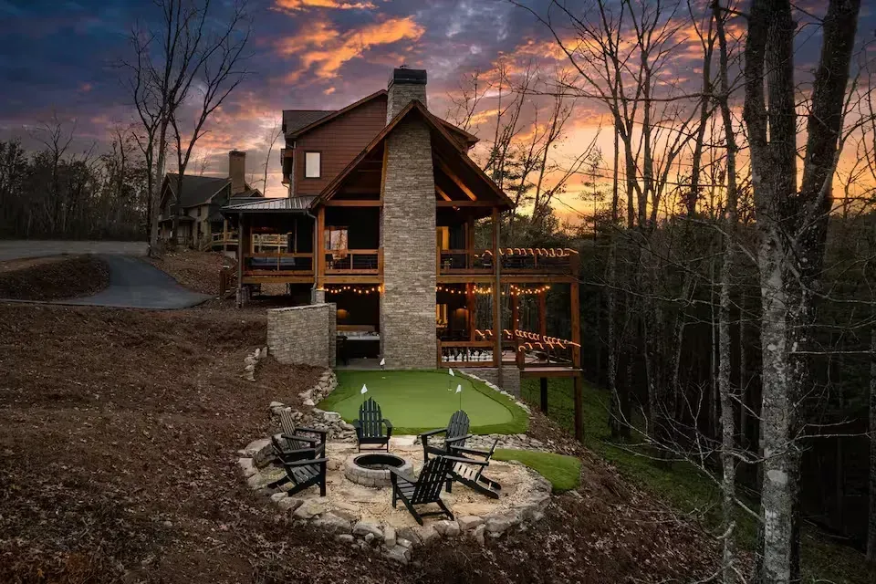 Rustic cabin at sunset with stone chimney, lit deck, and fire pit overlooking a wooded yard