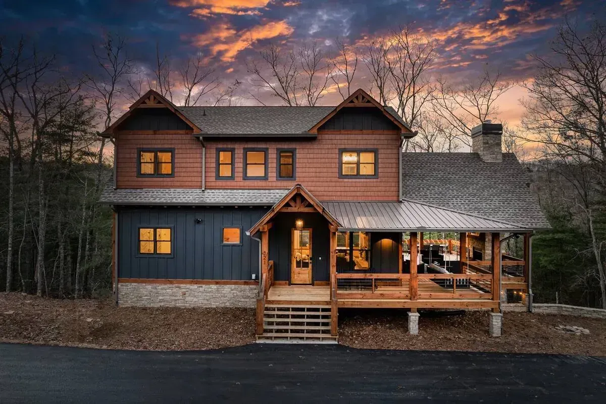 Rustic two-story cabin with lit windows and porch at dusk under a dramatic sunset sky