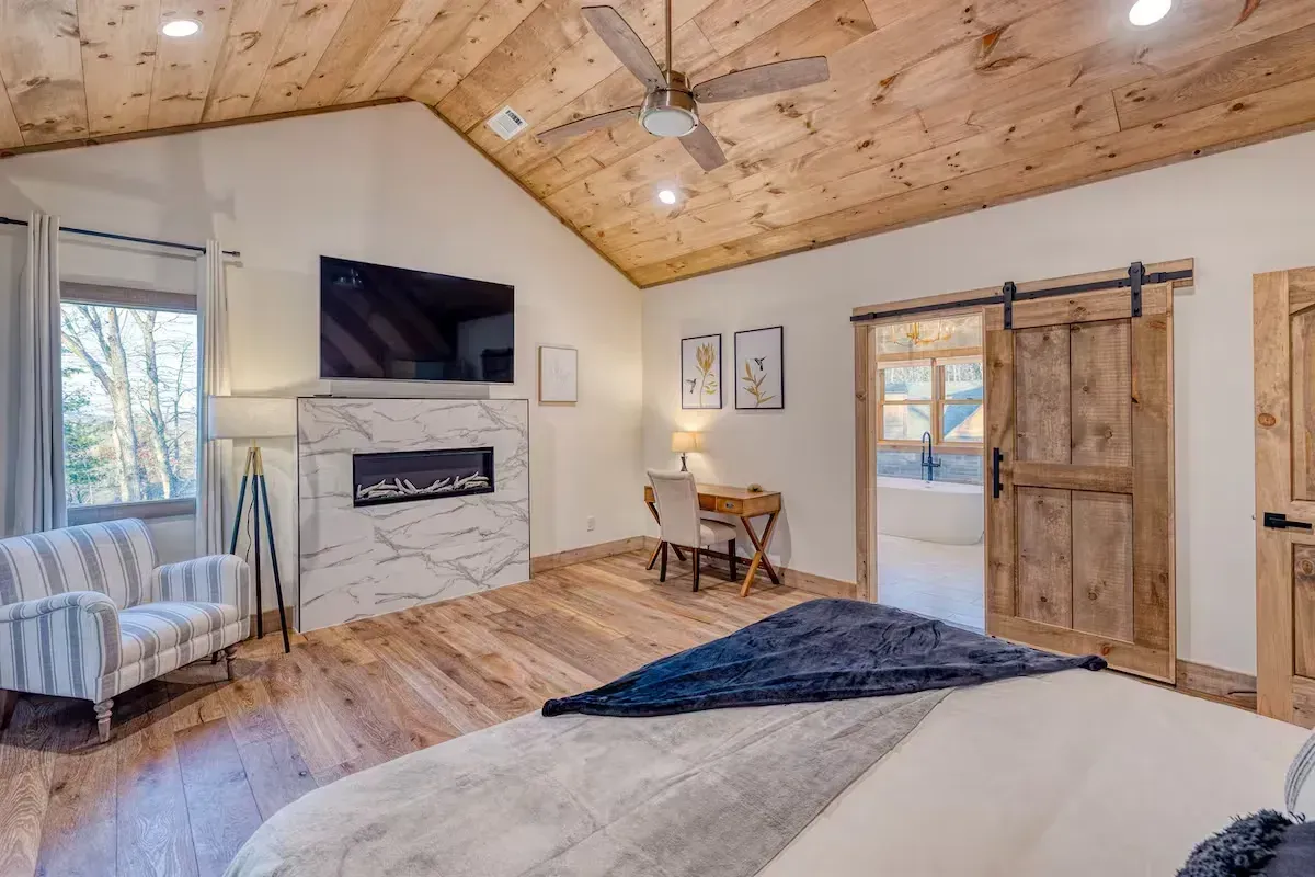 Cozy bedroom with vaulted wood ceiling, fireplace, wall-mounted TV, desk, and rustic sliding barn door.