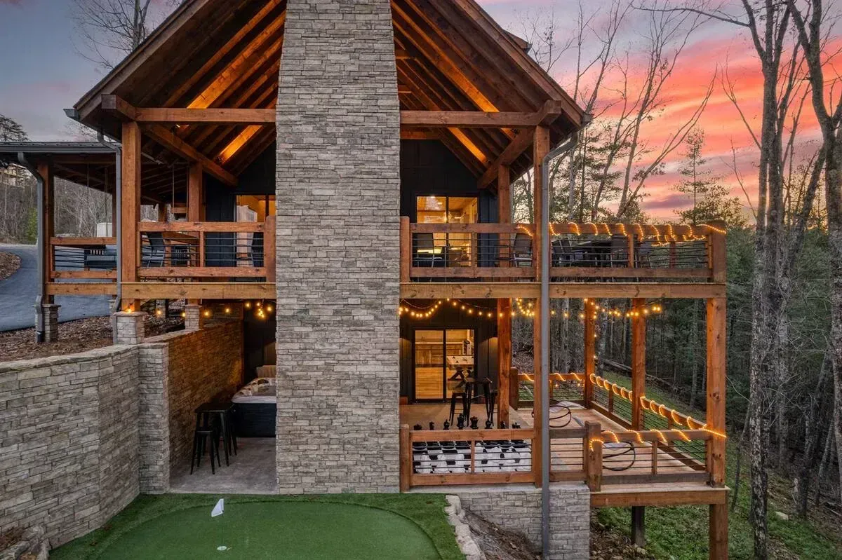 Rustic stone-and-wood cabin with balconies at sunset, surrounded by trees and a small lawn.