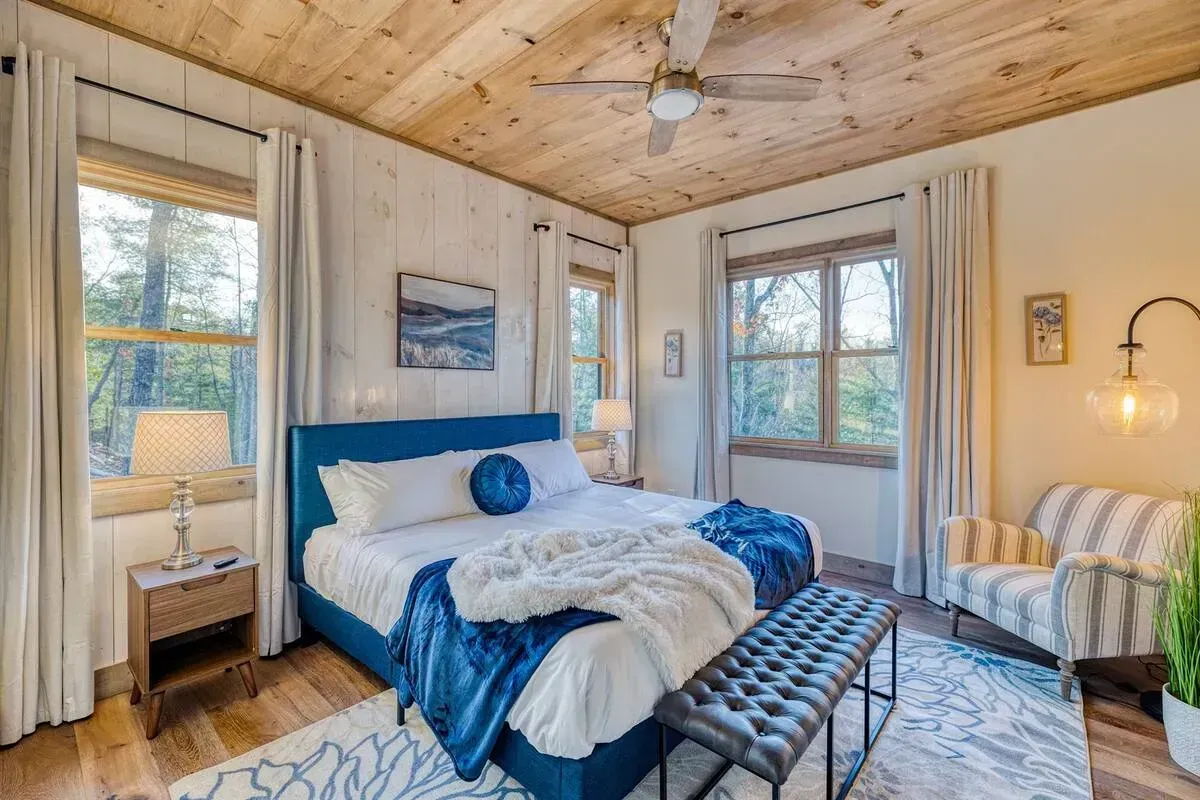Bright cozy bedroom with blue bedding, wood ceiling, two windows, and a bench at the foot of the bed