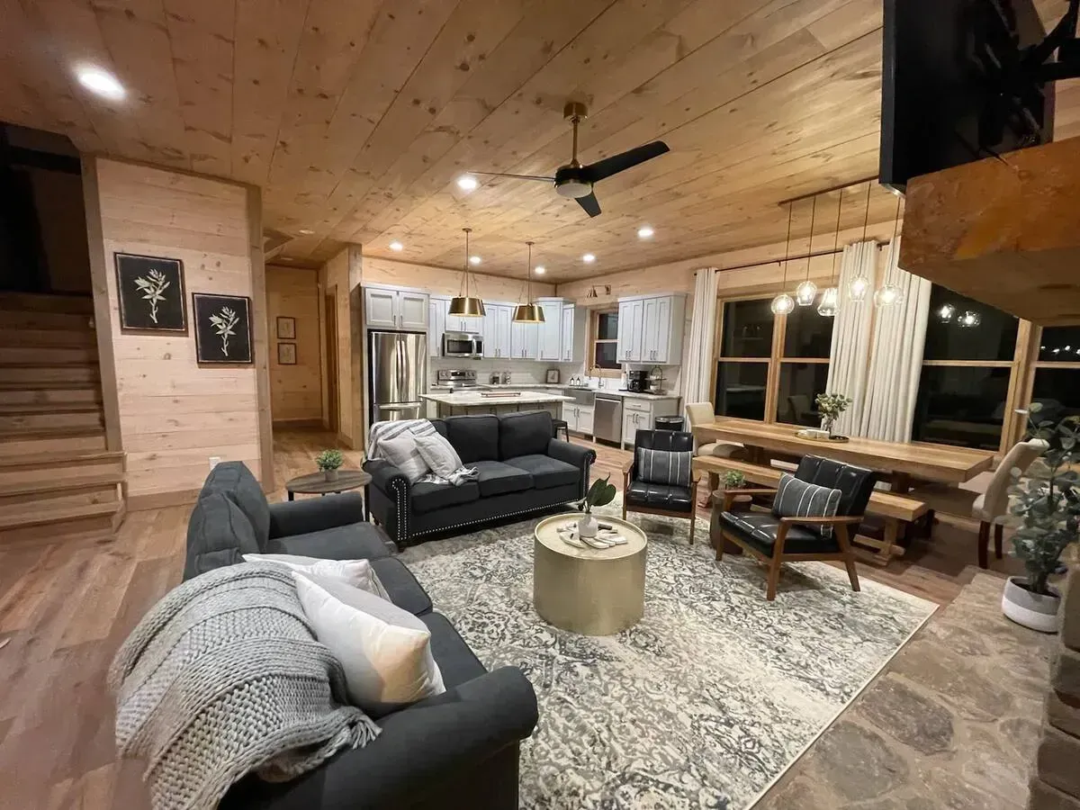 Cozy wood-paneled living room with gray sofas, rug, coffee table, and large windows at night
