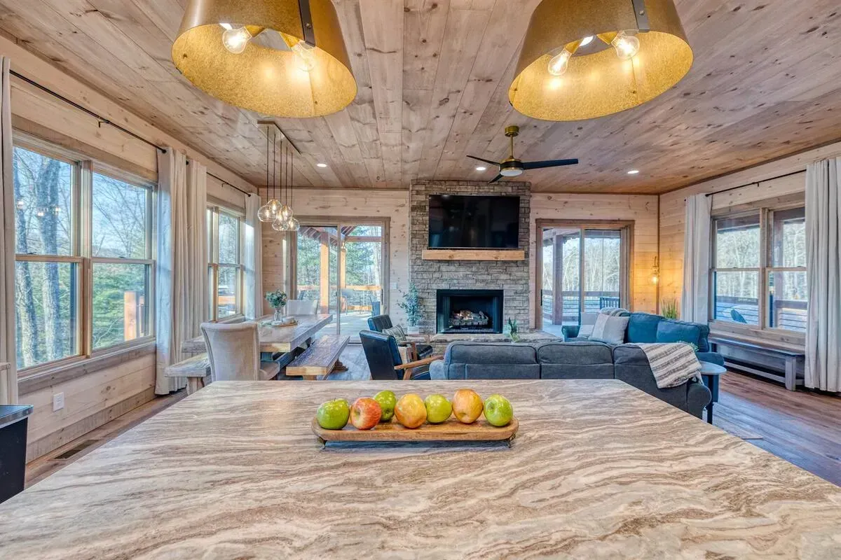 Cozy log cabin living room with stone fireplace, wood ceiling, large windows, and kitchen island in foreground