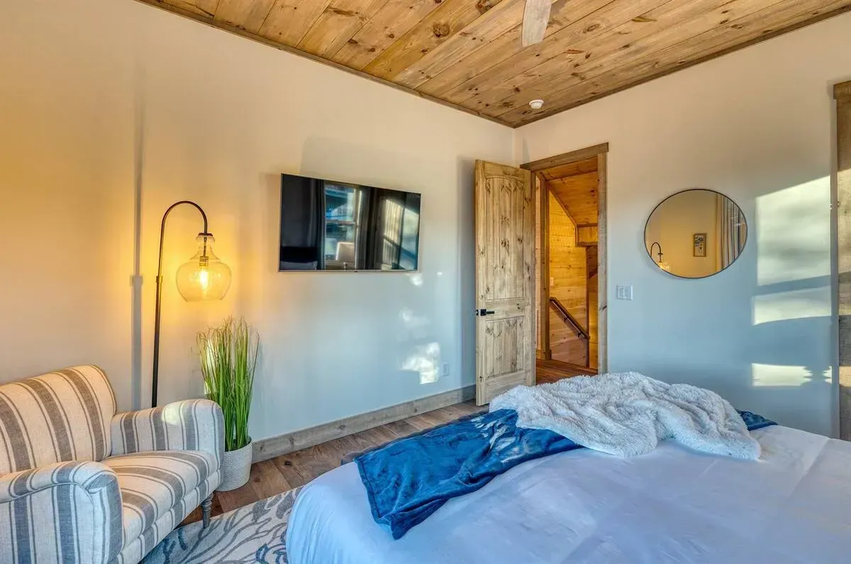 Cozy bedroom with wood ceiling, bed, striped chair, wall lamp, TV, and round mirror near a wooden door