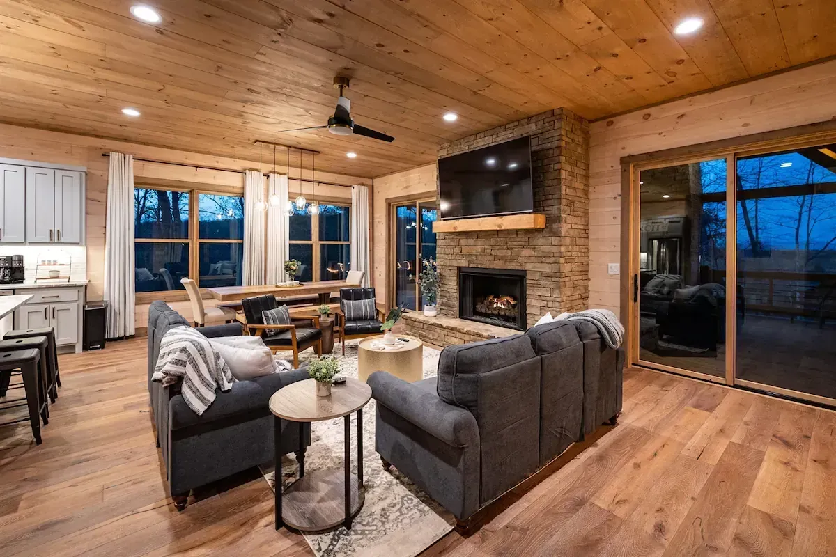 Cozy rustic living room with stone fireplace, wood ceiling, and gray sofas by large windows and glass doors