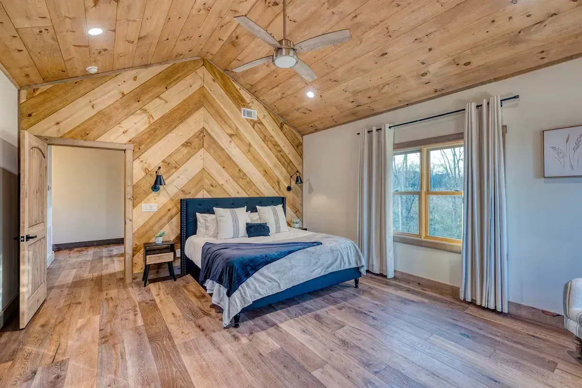 Spacious bedroom with wood-paneled ceiling, blue bedspread, chevron accent wall, and large windows