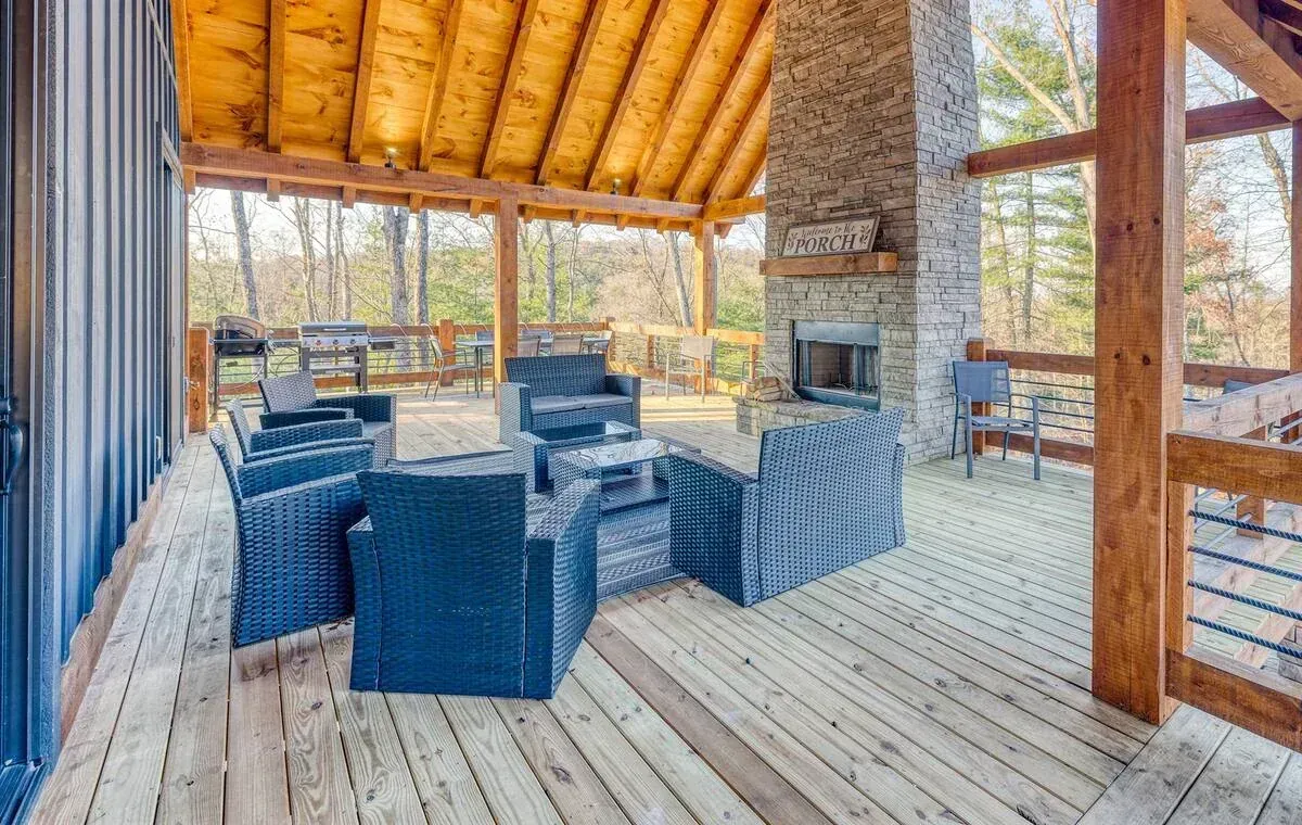 Covered wooden patio with blue wicker seating, a stone fireplace, and open views of trees.
