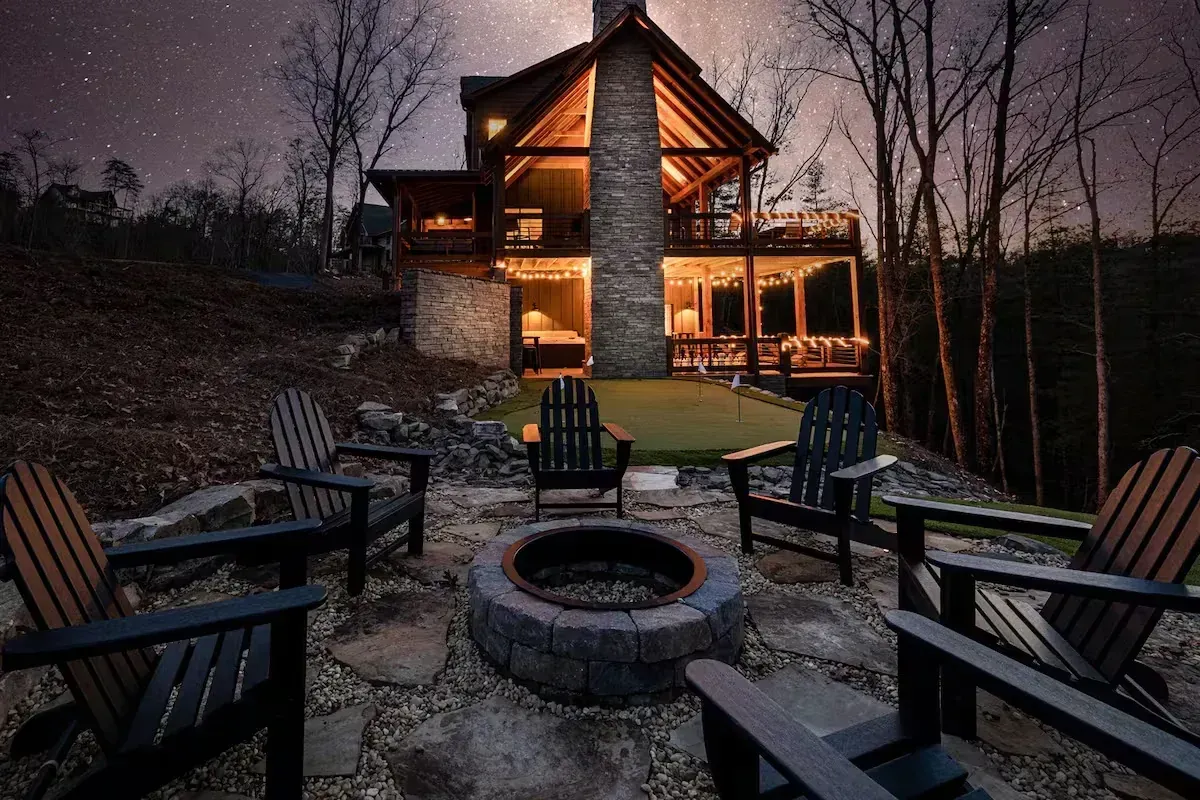 Cabin with lit windows at dusk, surrounded by Adirondack chairs around a stone fire pit.