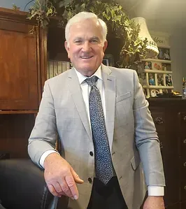 A man in a suit and tie is standing next to a chair in a room.