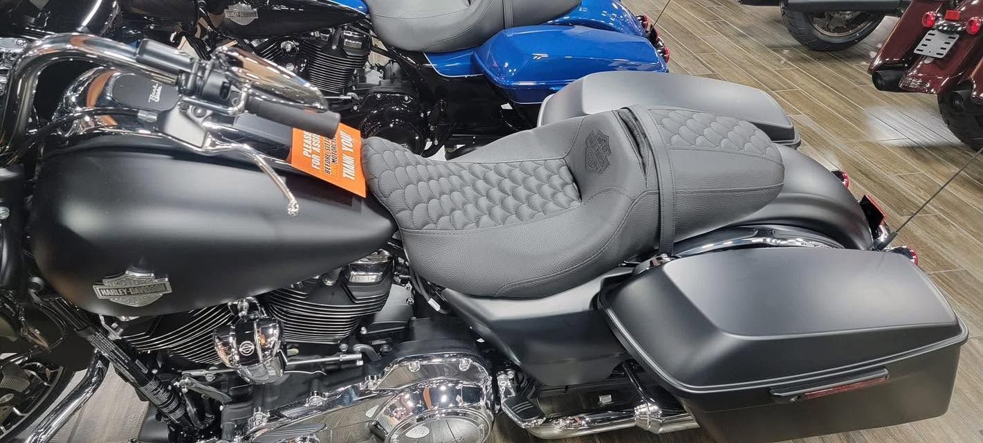 Black motorcycle with a gray quilted seat and saddlebags, parked indoors among other bikes