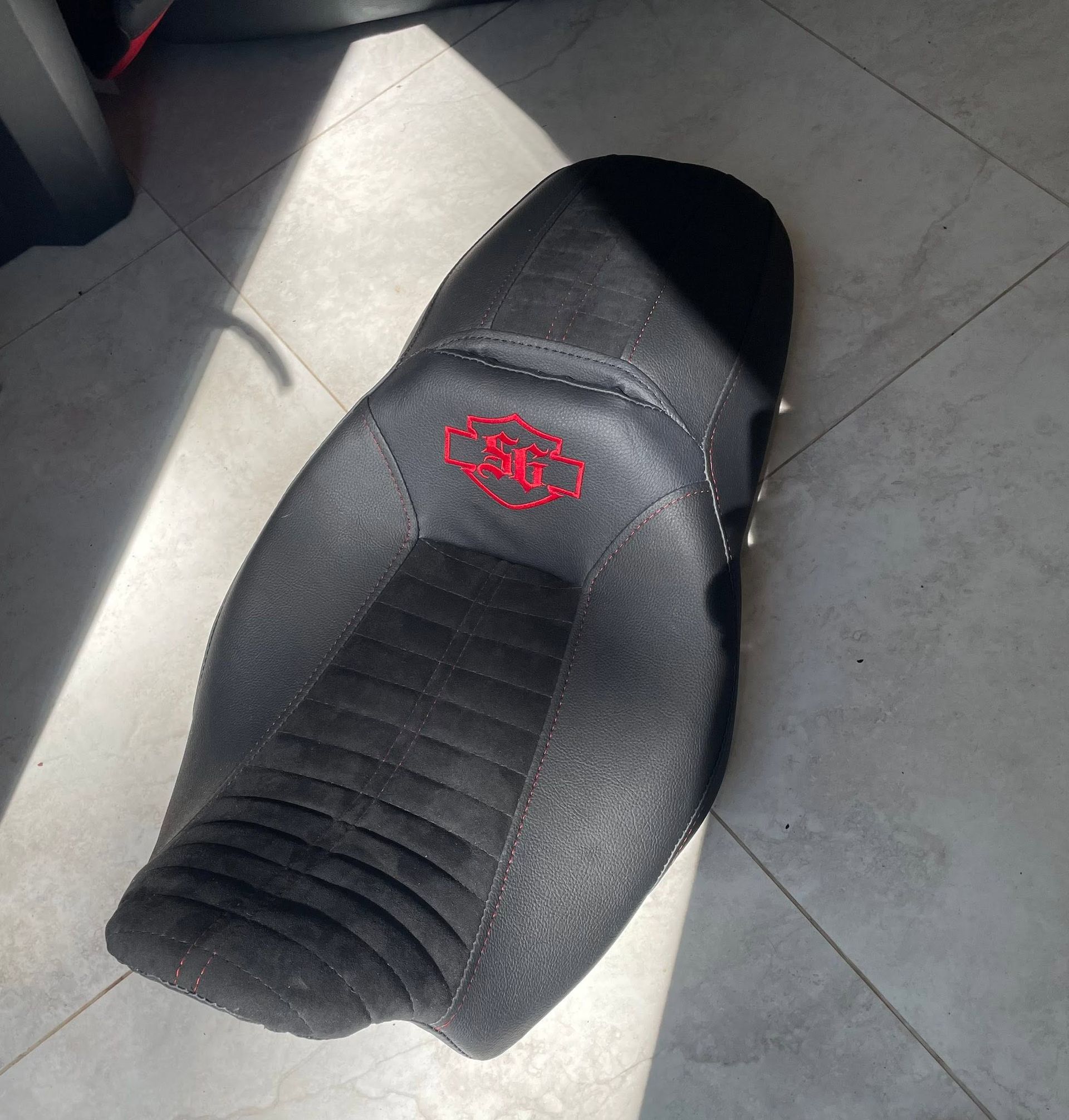 Black motorcycle seat on a tiled floor in sunlight, with a red embroidered logo.