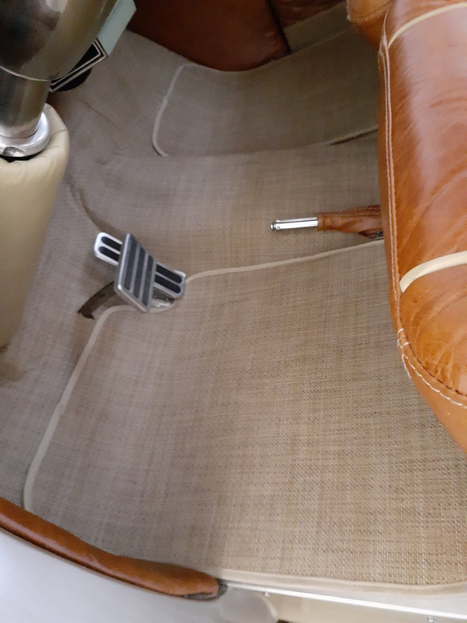 Close-up of a beige car seat with a metal pedal on the floor and a brown leather armrest