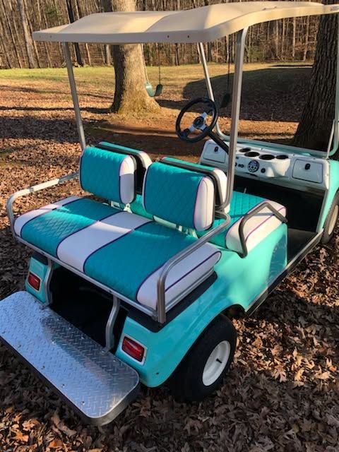 Teal golf cart with white seats and canopy parked on a leaf-covered path in a wooded area