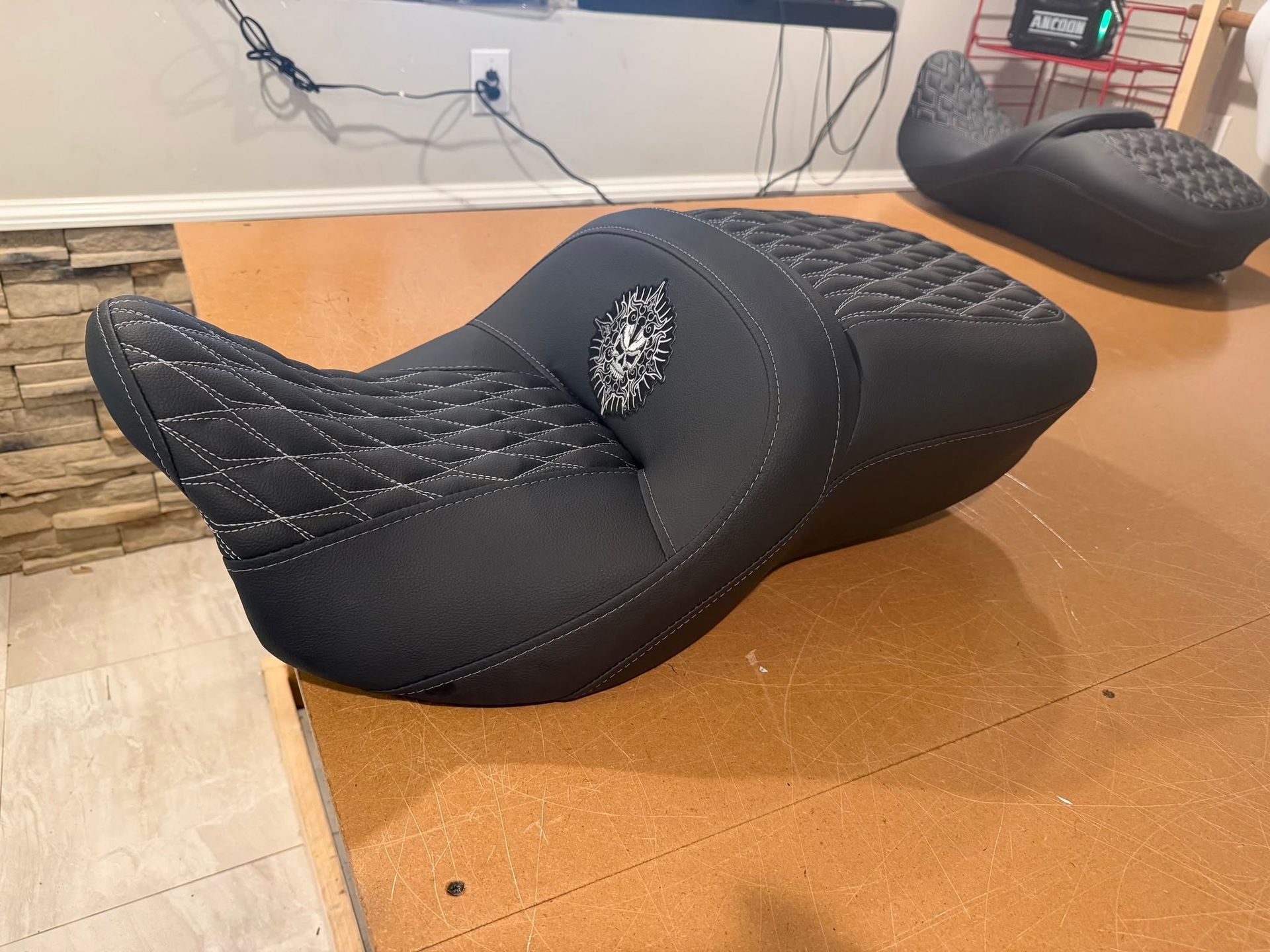 Black motorcycle seat on a wooden floor, with stitching and a center emblem.
