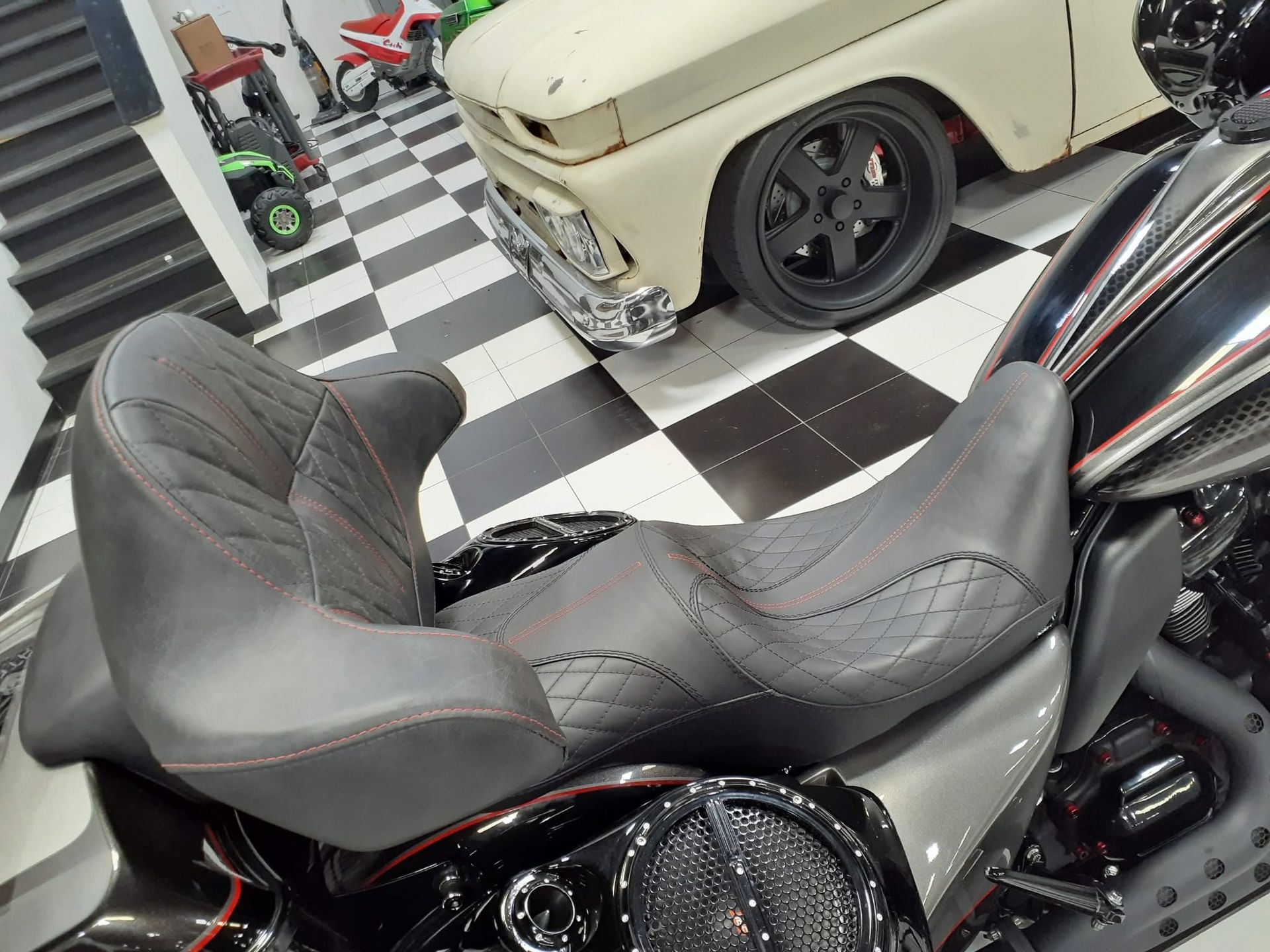 Black motorcycle in a garage with a checkered floor and a cream classic car in the background