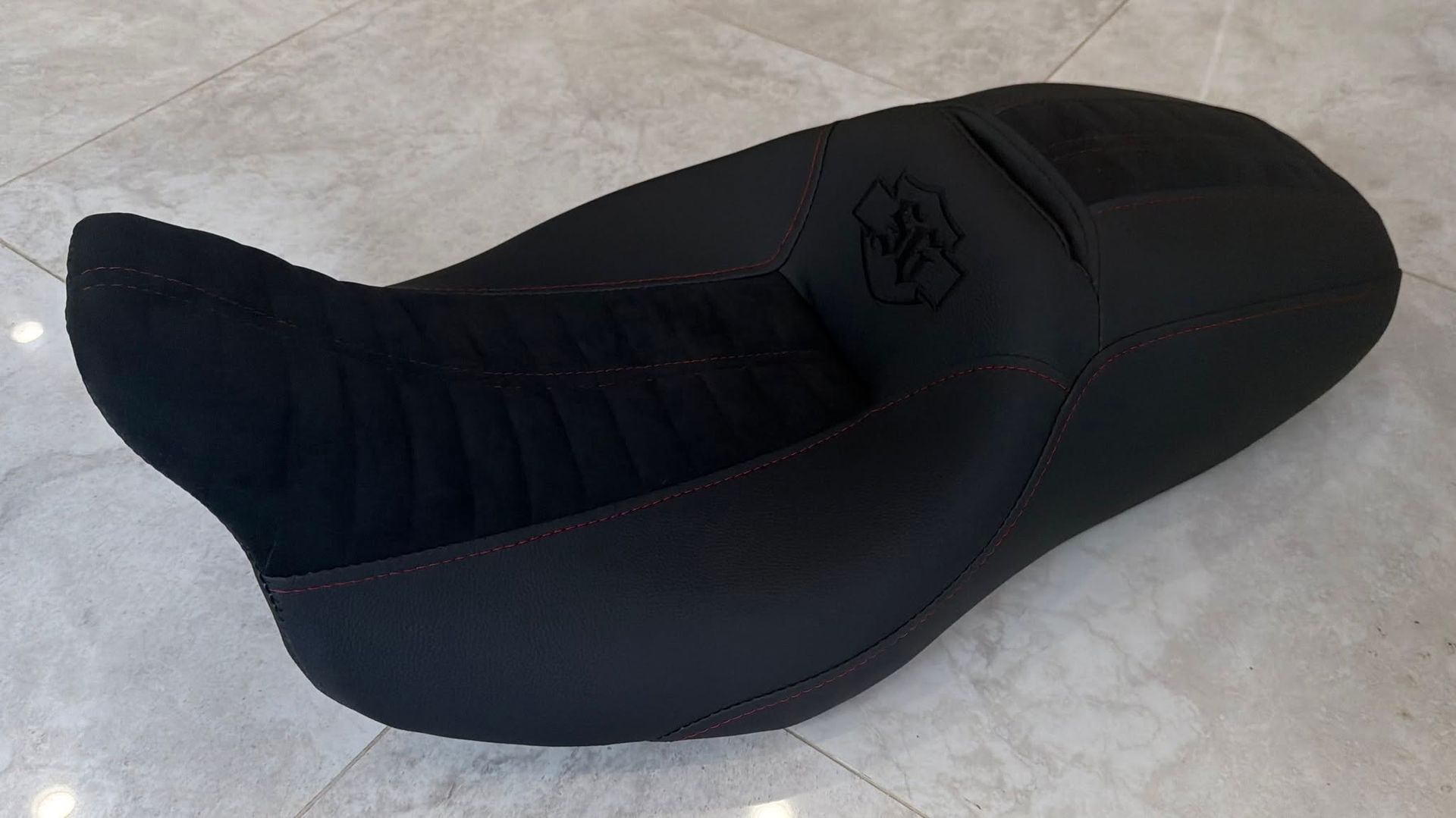 Black contoured motorcycle seat with white stitching and logo on a light tiled floor