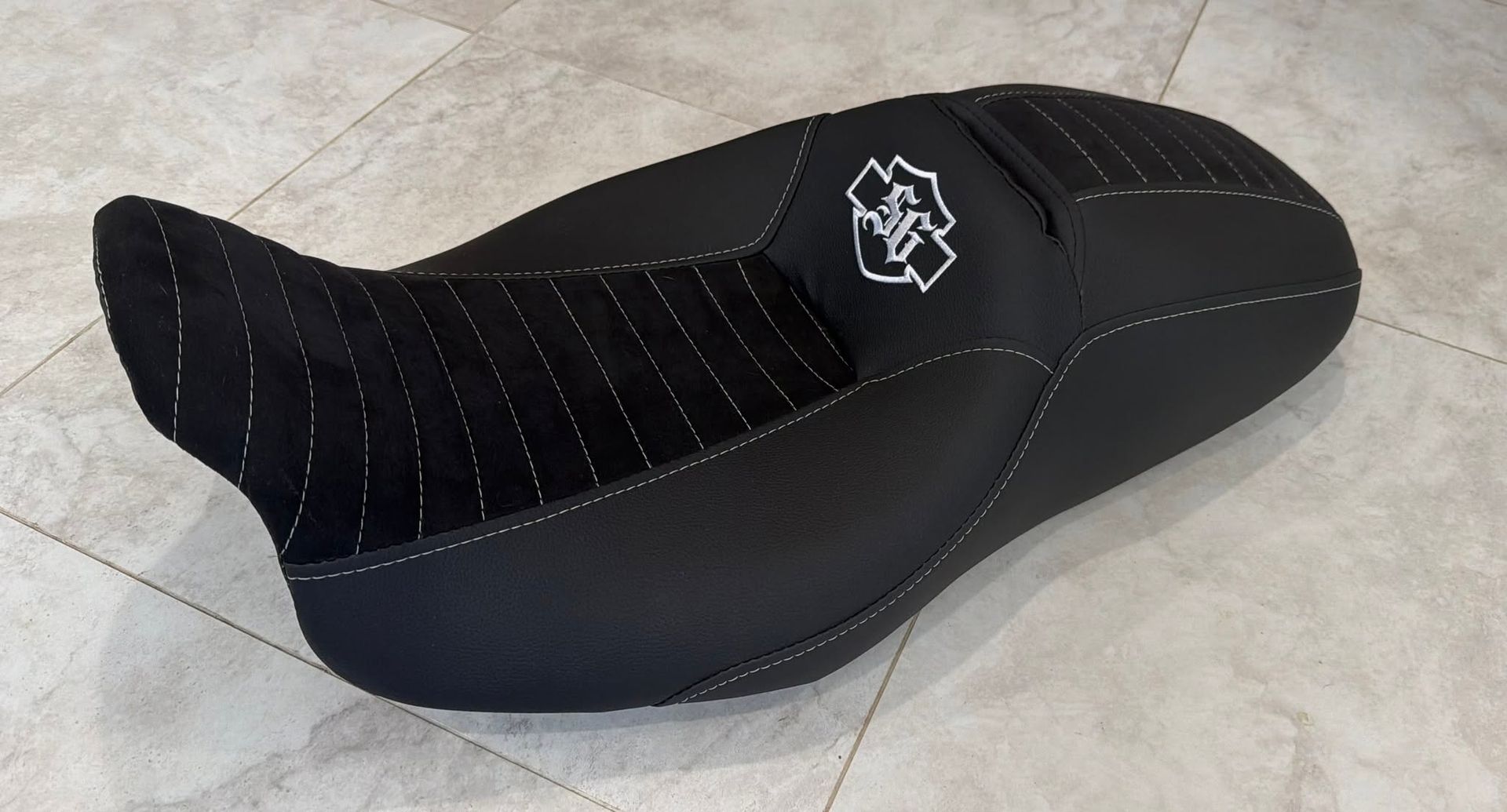 Black contoured motorcycle seat with white stitching and logo on a light tiled floor