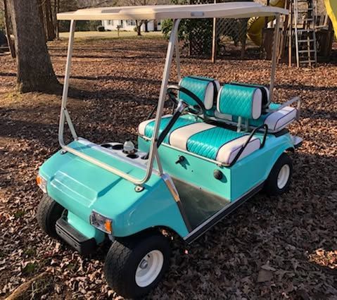 Turquoise golf cart with white roof parked on leaf-covered ground under trees