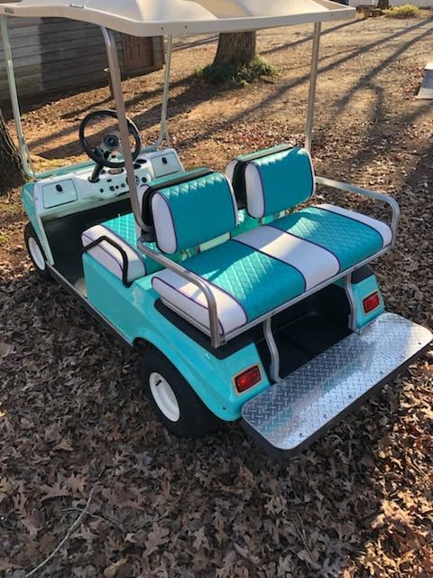 Turquoise golf cart with white roof and bench seats parked on a leaf-covered path.