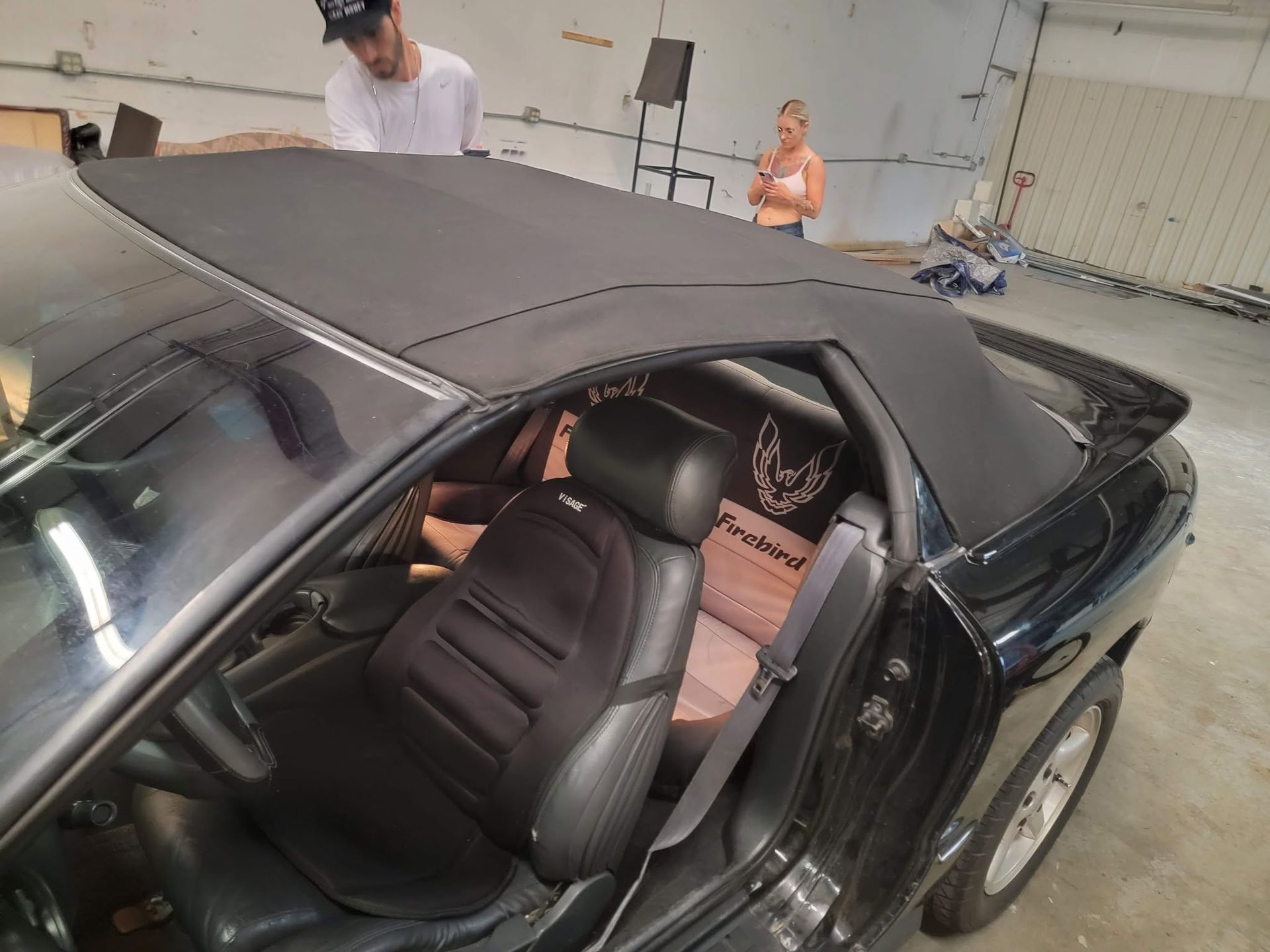 Black convertible with its soft top being installed in a workshop, interior seats visible.