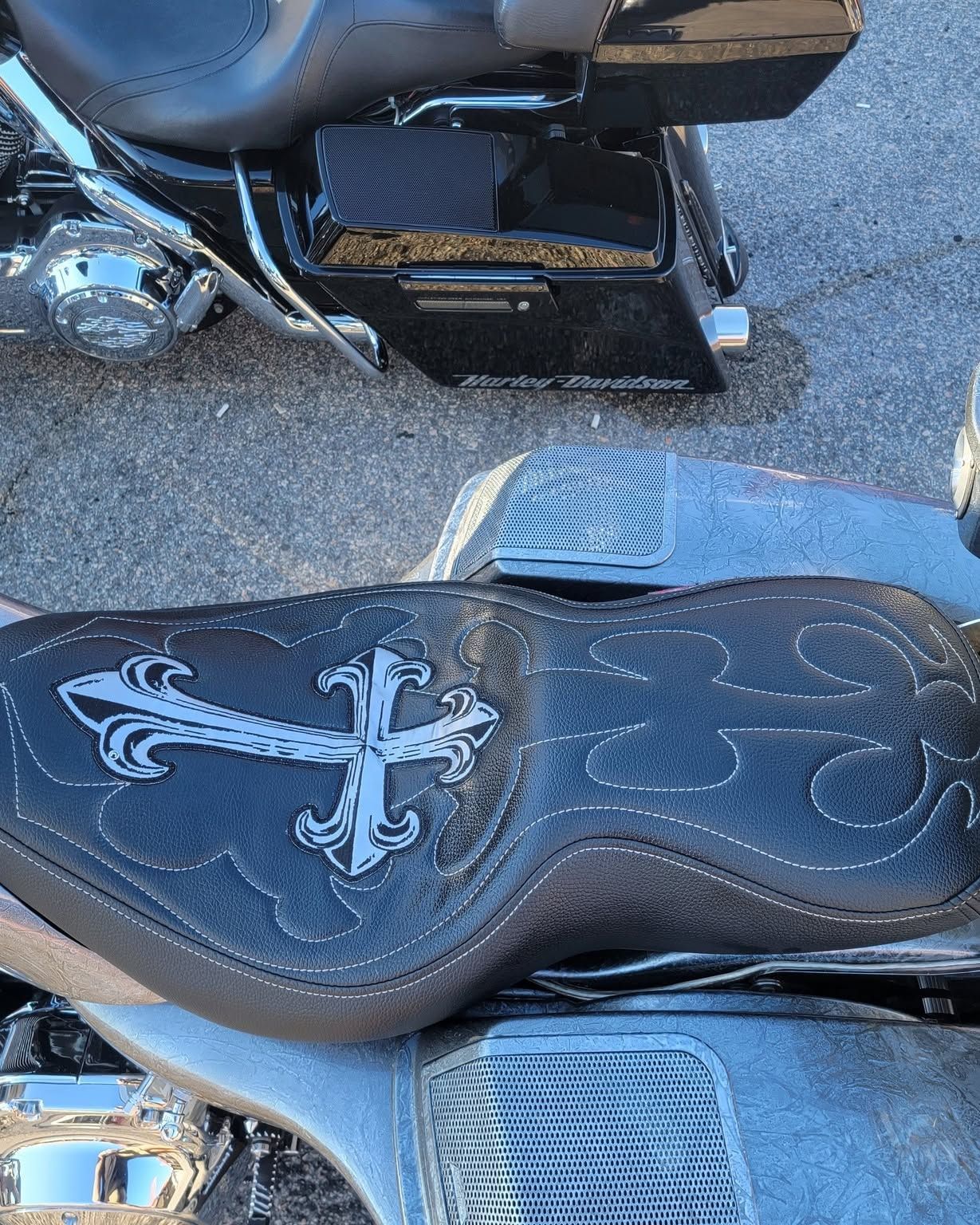 Close-up of a black motorcycle seat with a silver cross emblem and footboards on asphalt.