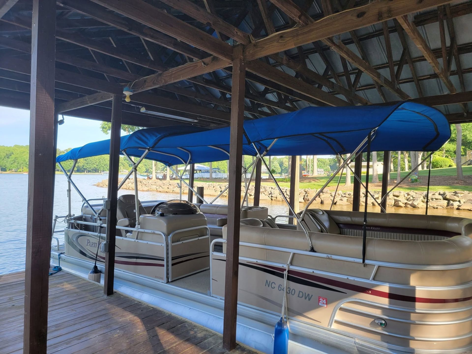 Covered pontoon boat docked under a wooden boathouse, with blue canopy and lake in the background