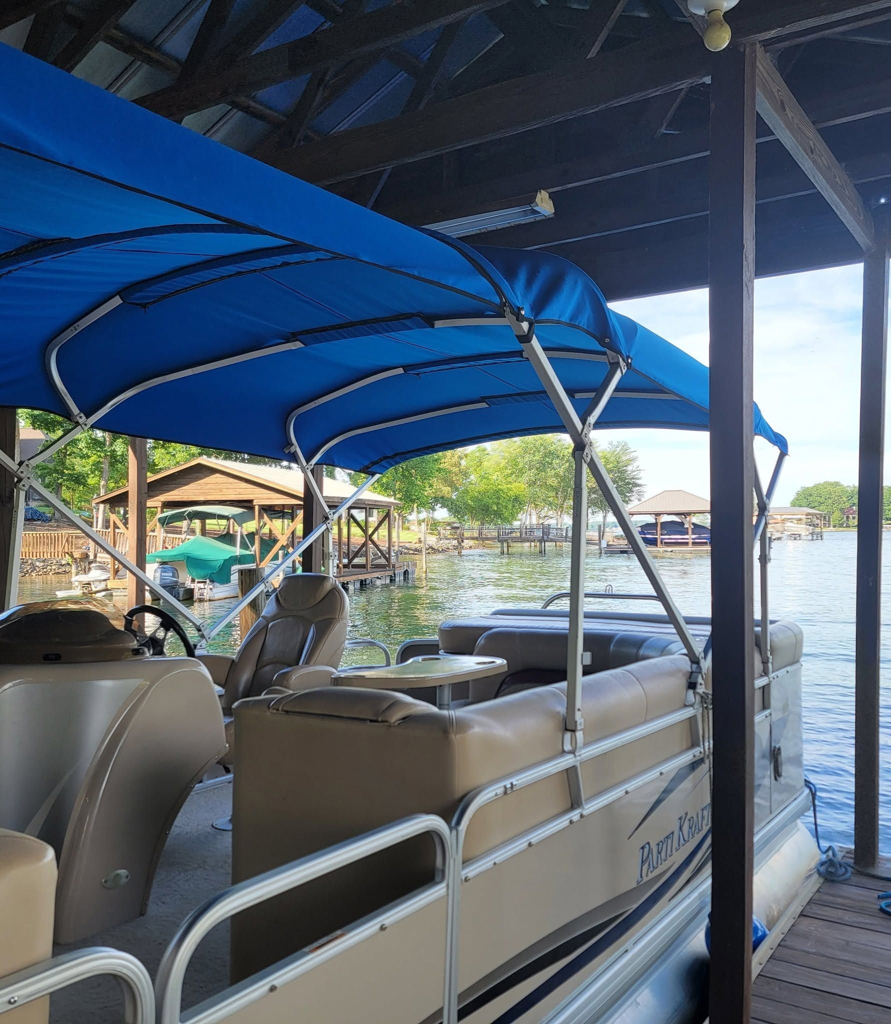 Blue-canopy pontoon boat docked beside a lakeside pier, with water and shoreline in the background