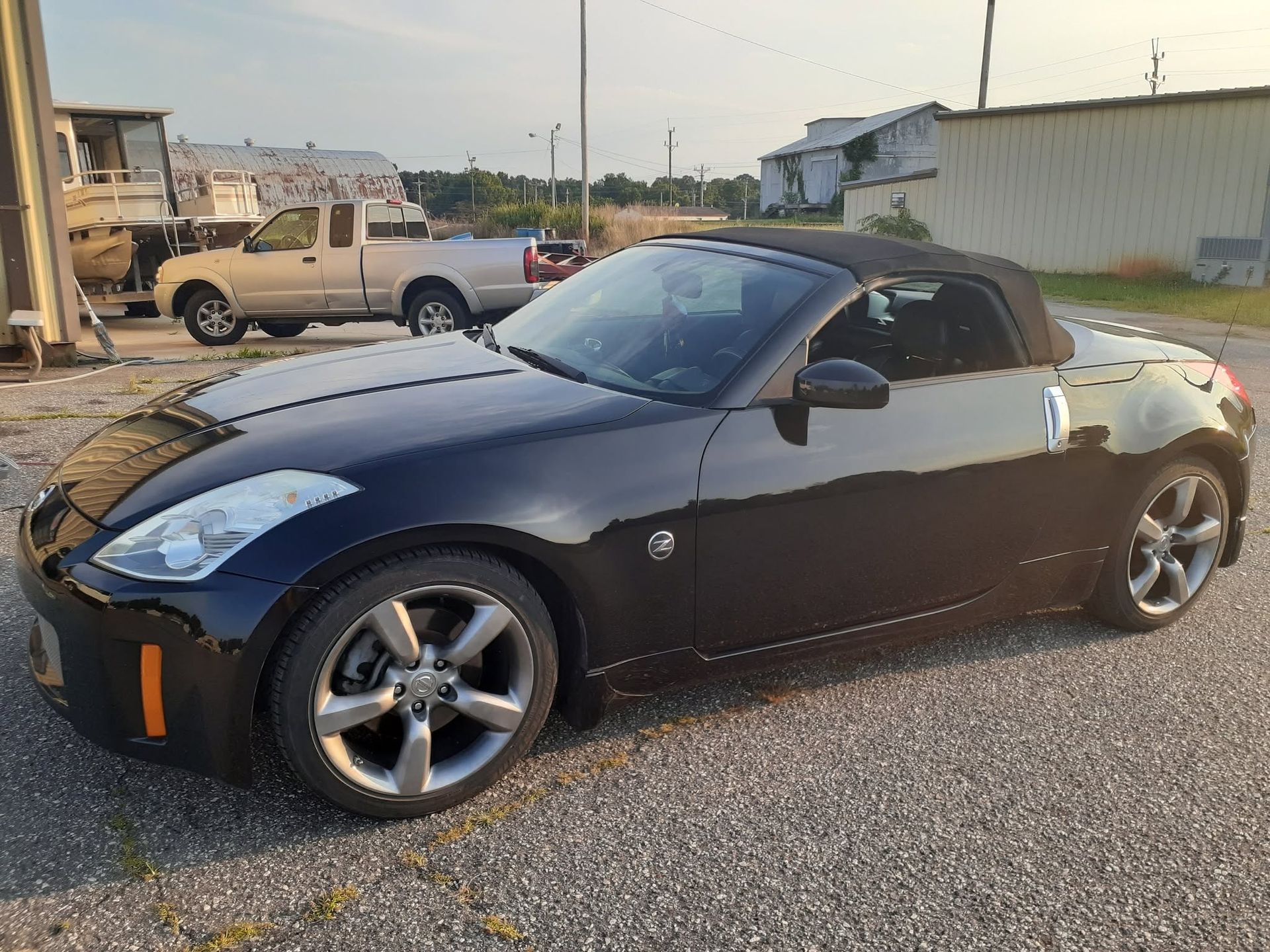 Black Nissan 350Z convertible parked on gravel near industrial buildings at dusk