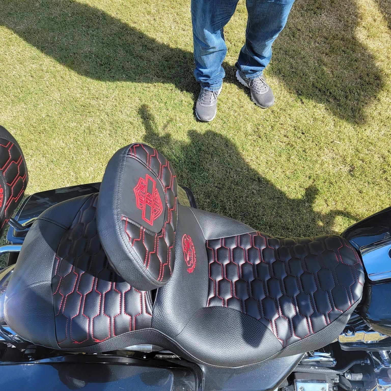 Black saddle with red stitching on a motorcycle, viewed from above on grass with a person's feet nearby