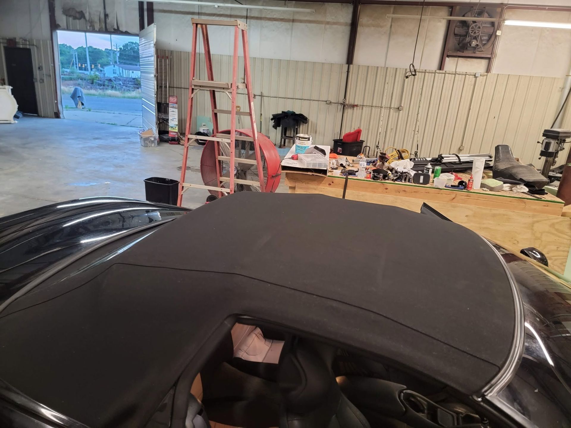 Black convertible top in a garage workshop with ladders, tools, and a workbench in the background