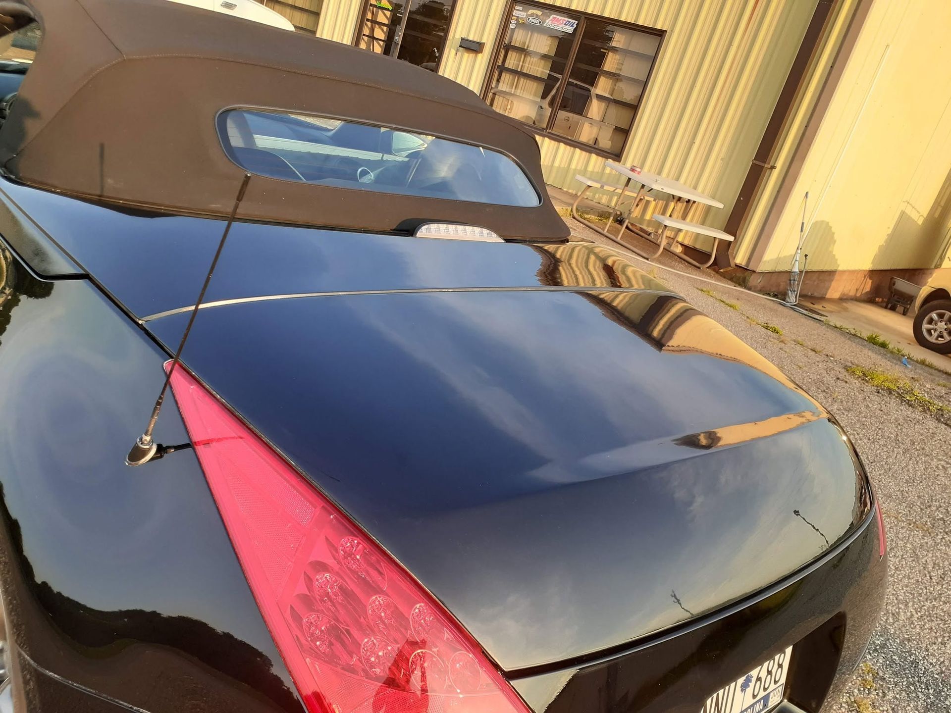 Black sports car with brown soft top parked outdoors near a building and gravel driveway