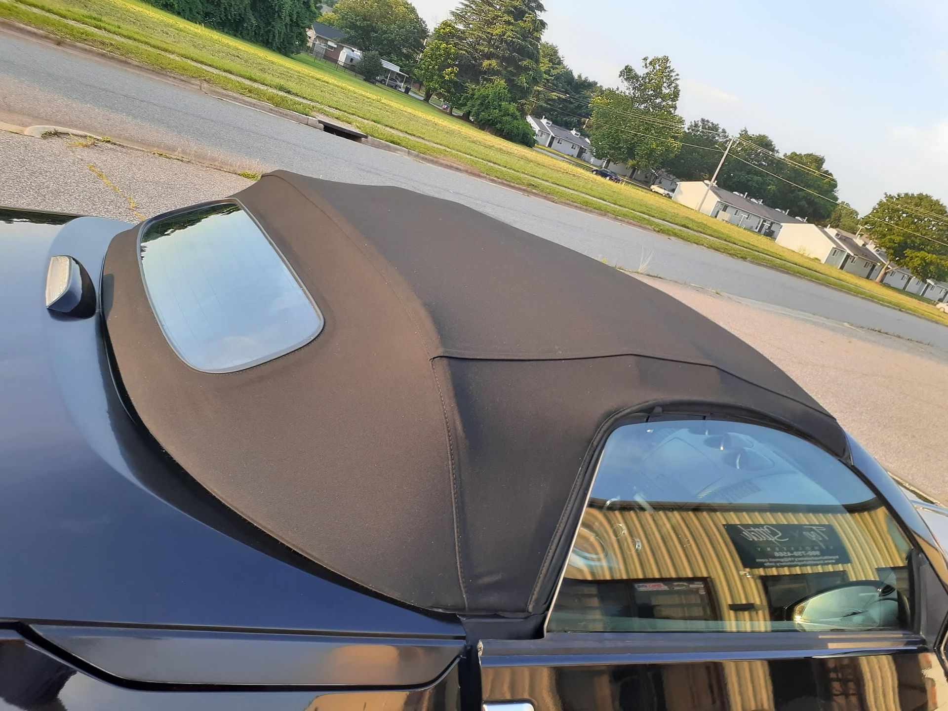 Black convertible with a brown soft top and rear window, parked on a street near houses.