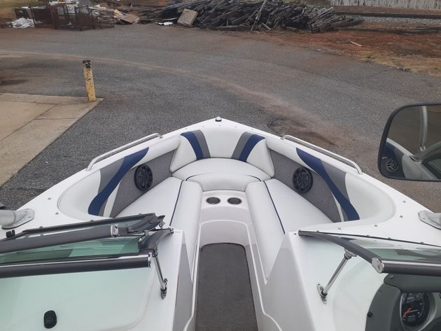 Front of a white-and-blue boat on a gravel lot, viewed from the cockpit with windshield and steering wheel visible