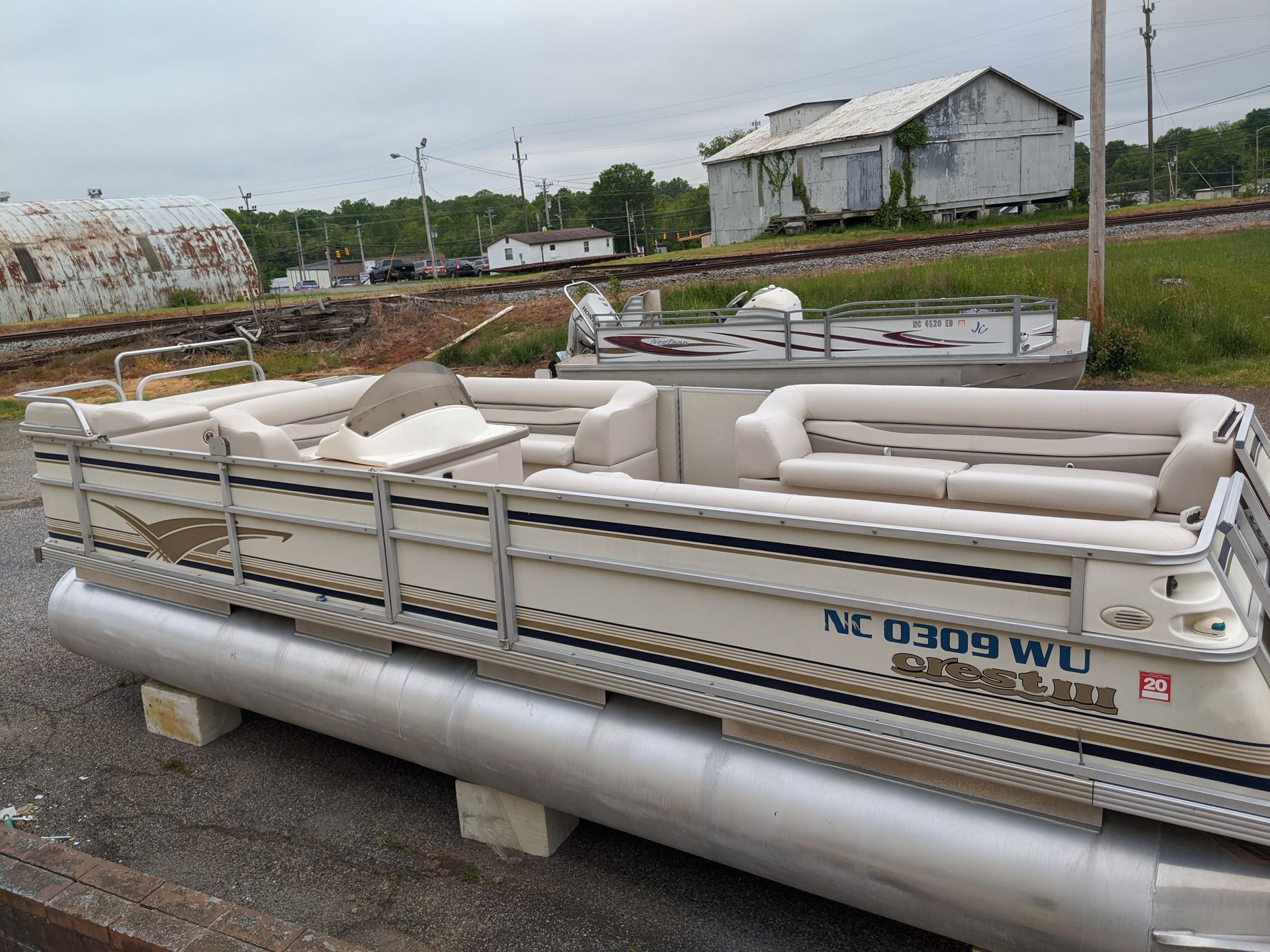 White pontoon boat on a trailer in a rural yard with houses in the background