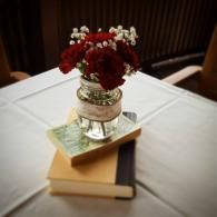 Small red floral arrangement in a silver vase atop stacked books on a white tablecloth