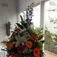 Large colorful flower arrangement with orange, purple, and white blooms in a bright indoor room by windows