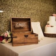 Wooden card box labeled “CARDS” on a wedding table beside a white tiered cake and flowers