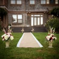 Wedding aisle on a grassy lawn leading to a stone house, lined with white fabric and floral arrangements