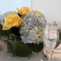 Yellow roses and pale blue hydrangeas in a glass vase beside a water goblet on a white table